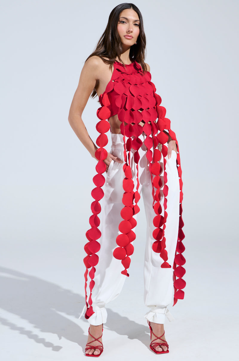 A woman poses in the LIGHTS FLASHING HALTER TIE NECK TOP IN RED with white pants and red high-heeled sandals, standing against a plain light background. The top features large round fabric pieces hanging in strips.