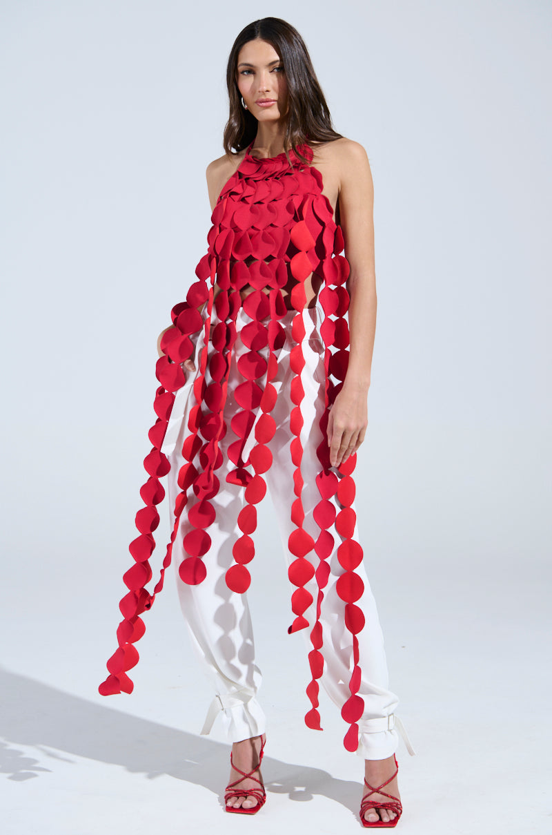 A woman poses confidently in white pants and red heels, wearing the LIGHTS FLASHING HALTER TIE NECK TOP IN RED—a unique top featuring long, hanging red circular shapes that drape from her neck to her legs.