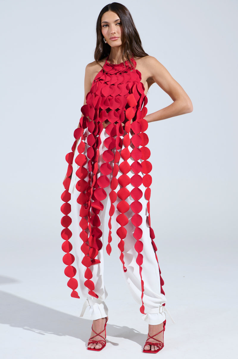 A woman in red high heels and white pants confidently wears the LIGHTS FLASHING HALTER TIE NECK TOP IN RED, featuring a sleeveless design with long, hanging strips of circular fabric shapes, against a plain light background.