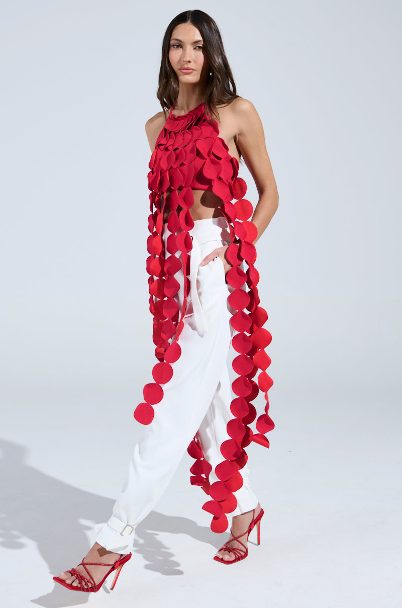 A woman showcases the LIGHTS FLASHING HALTER TIE NECK TOP IN RED, a sleeveless piece with large circular appliqués on strands, styled with loose white pants and red strappy heels against a plain light background.
