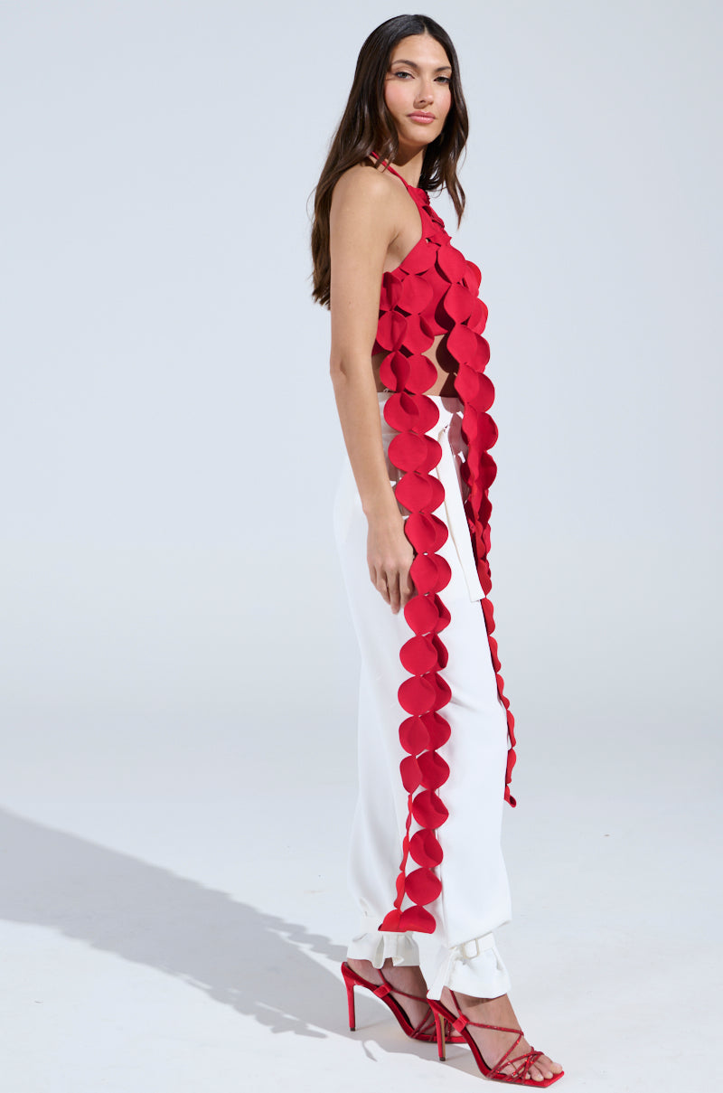 A woman stands sideways wearing the LIGHTS FLASHING HALTER TIE NECK TOP IN RED, paired with loose white ruffled-hem pants and red strappy heels, set against a plain light background.