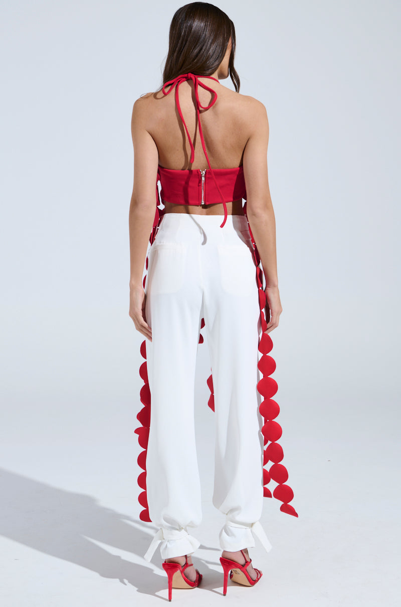 A woman faces away, wearing the LIGHTS FLASHING HALTER TIE NECK TOP IN RED with a metallic back zipper, paired with white high-waisted pants featuring red circular appliqués on the sides and red high heels for a bold look.