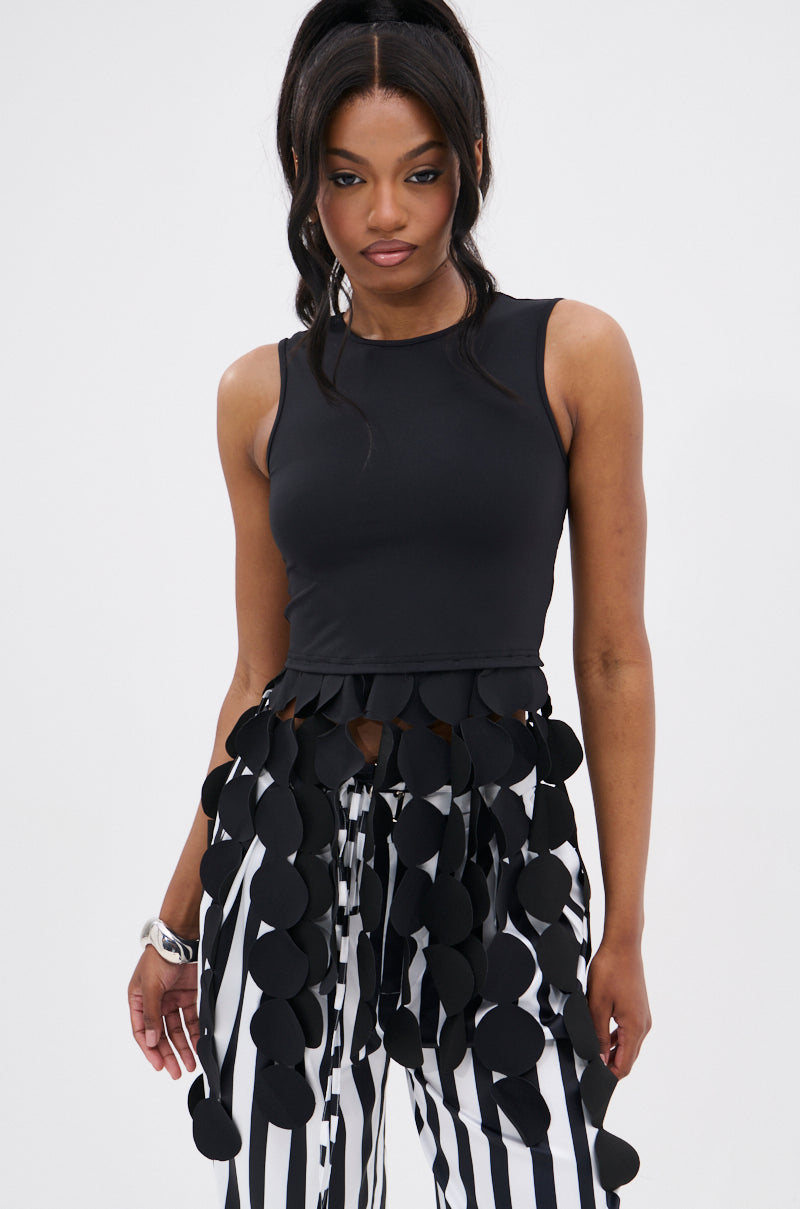 A woman wears the LIGHTS FLASHING TOP, a sleeveless black bodycon with circular cut-outs at the hem, paired with high-waisted black-and-white vertical striped pants. Her long, wavy hair is styled in a high ponytail.