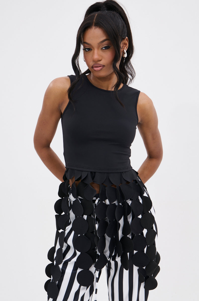 A woman poses against a plain background in the LIGHTS FLASHING TOP, a sleeveless black top with disc-link fringe at the hem, styled with vertically striped black-and-white pants featuring oversized black circular appliqués.