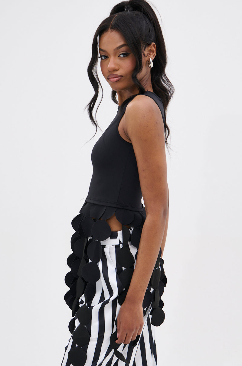 A woman with long, wavy hair in a high ponytail wears the LIGHTS FLASHING TOP with disc-link fringe and high-waisted black and white vertical striped pants, posing against a plain white background.