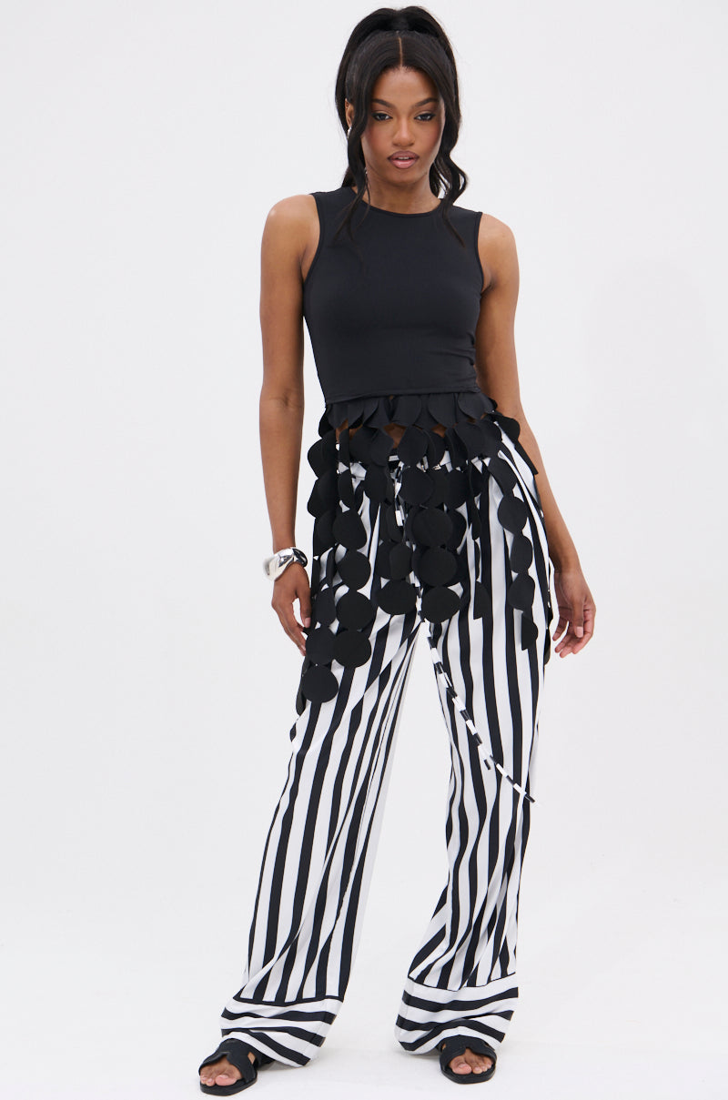 A woman stands against a plain white background wearing the LIGHTS FLASHING TOP, a sleeveless black top with large circular accents, paired with high-waisted black and white vertical striped wide-leg pants.