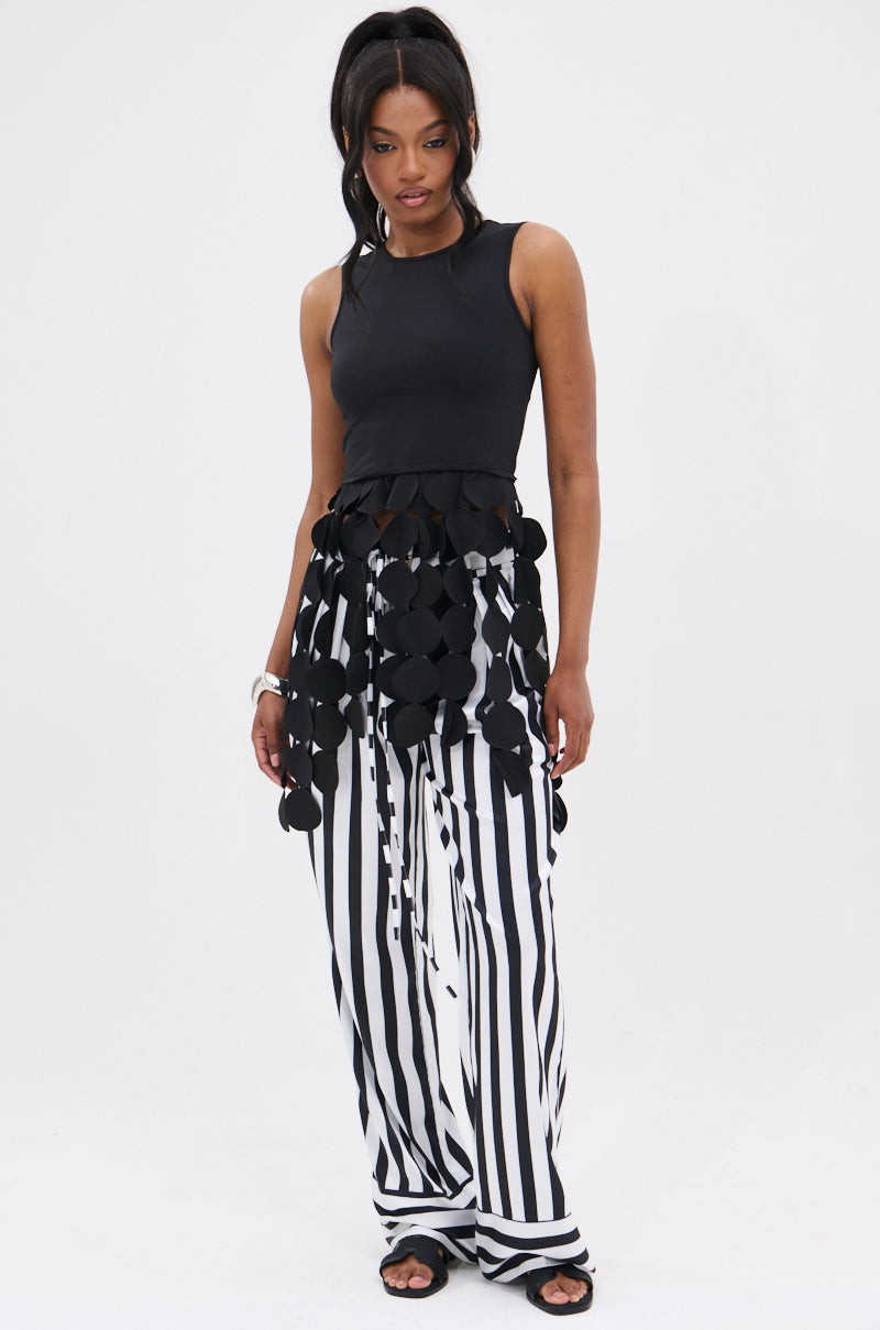 A woman stands against a white background wearing the LIGHTS FLASHING TOP—sleeveless, black, with circular appliqués on sheer fabric—paired with wide-leg black and white striped pants and black sandals.