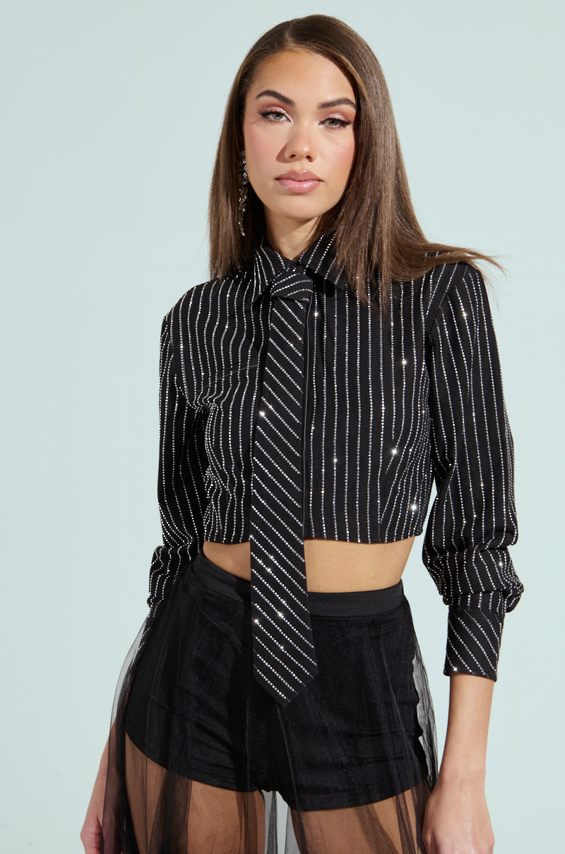 A person is wearing the "JUST GO WITH IT BUTTON DOWN TOP WITH TIE," featuring black and white stripes in a long-sleeve, cropped, boxy silhouette. They pair it with black shorts with a sheer overlay. The backdrop is a plain light blue.
