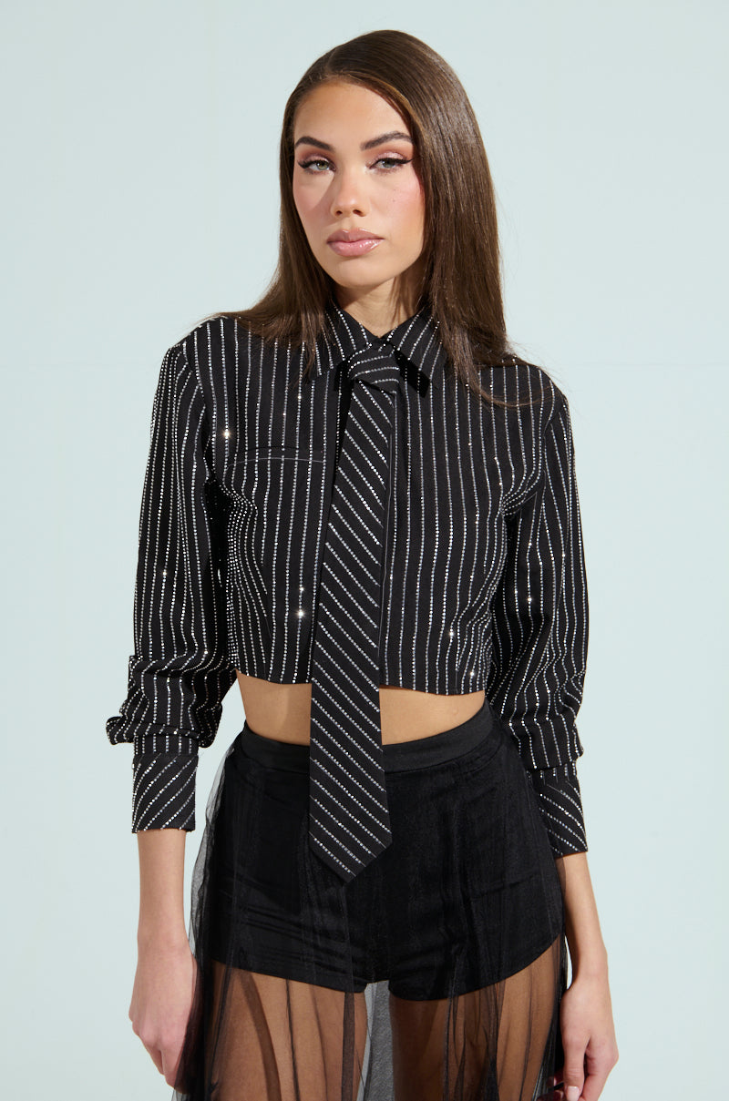 A person with long brown hair wears the "Just Go With It Button Down Top with Tie," a black cropped shirt featuring a rhinestone pinstripe and matching tie. They pair it with sheer black shorts over black bottoms against a plain light gray background, enhancing the outfit’s modern appeal.