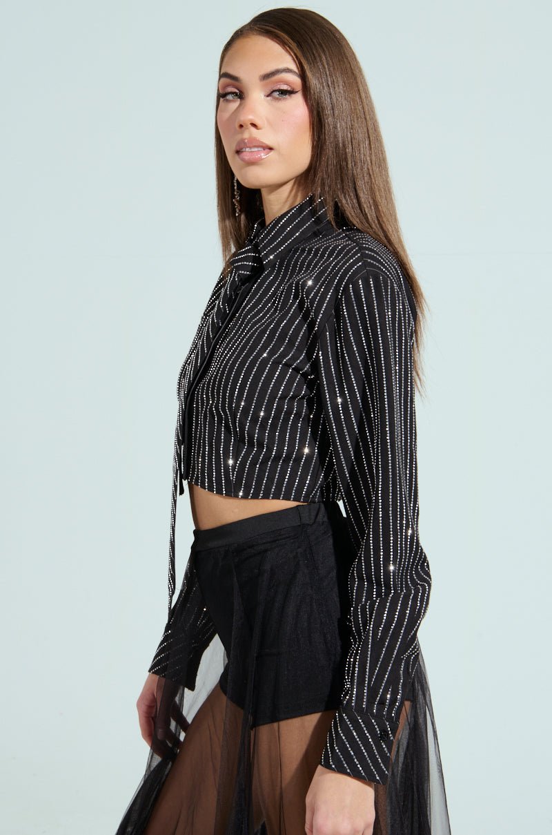 In a light blue setting, a woman with long brown hair elegantly poses in the JUST GO WITH IT BUTTON DOWN TOP WITH TIE. Her gaze is directed to the side, highlighting the black striped sequin details and rhinestone pinstripe pattern on its sheer overlay.