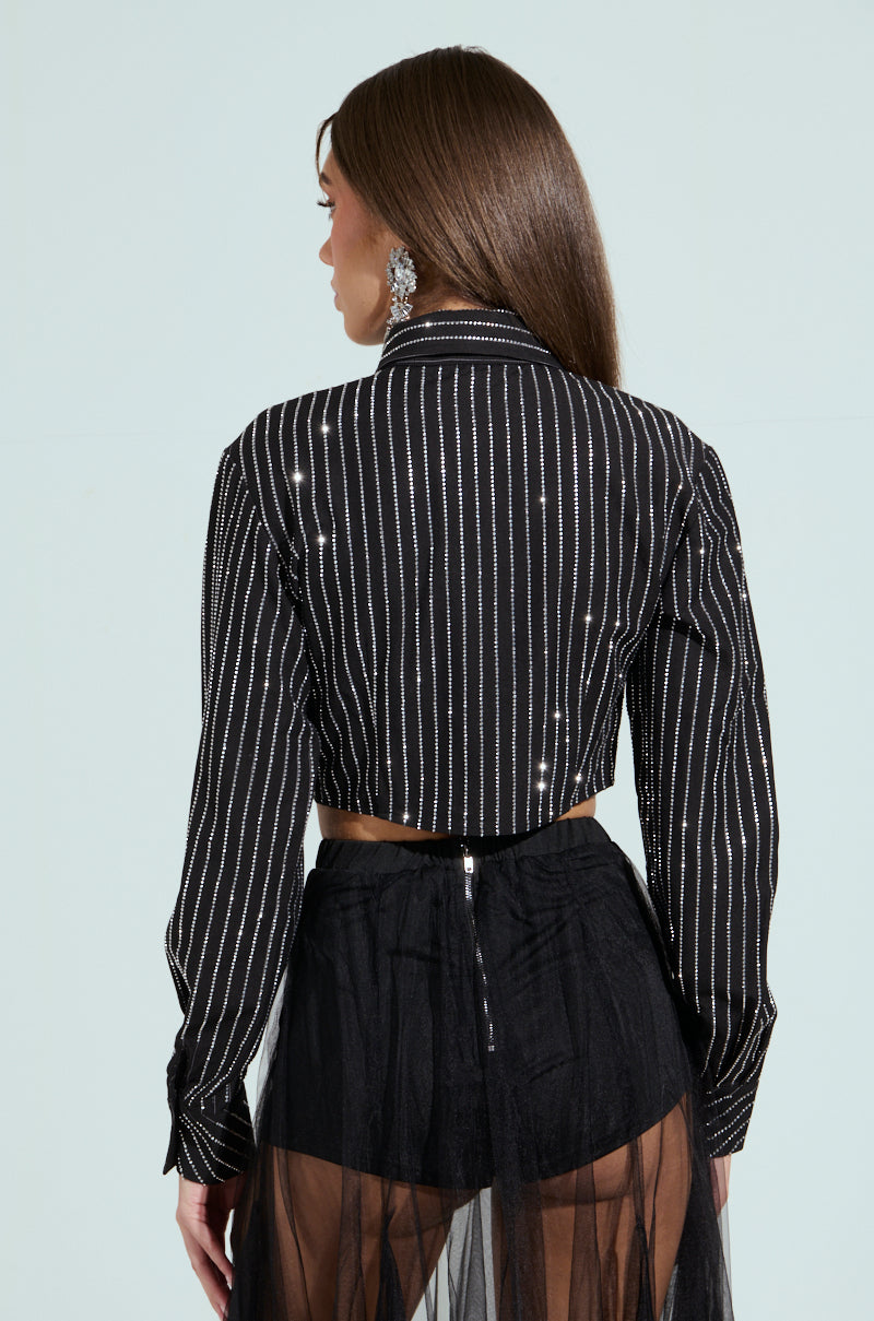 A woman with long brown hair is shown from behind, wearing the JUST GO WITH IT BUTTON DOWN TOP WITH TIE, ornate earrings, and a sheer black skirt over high-waisted black shorts, standing against a light background.