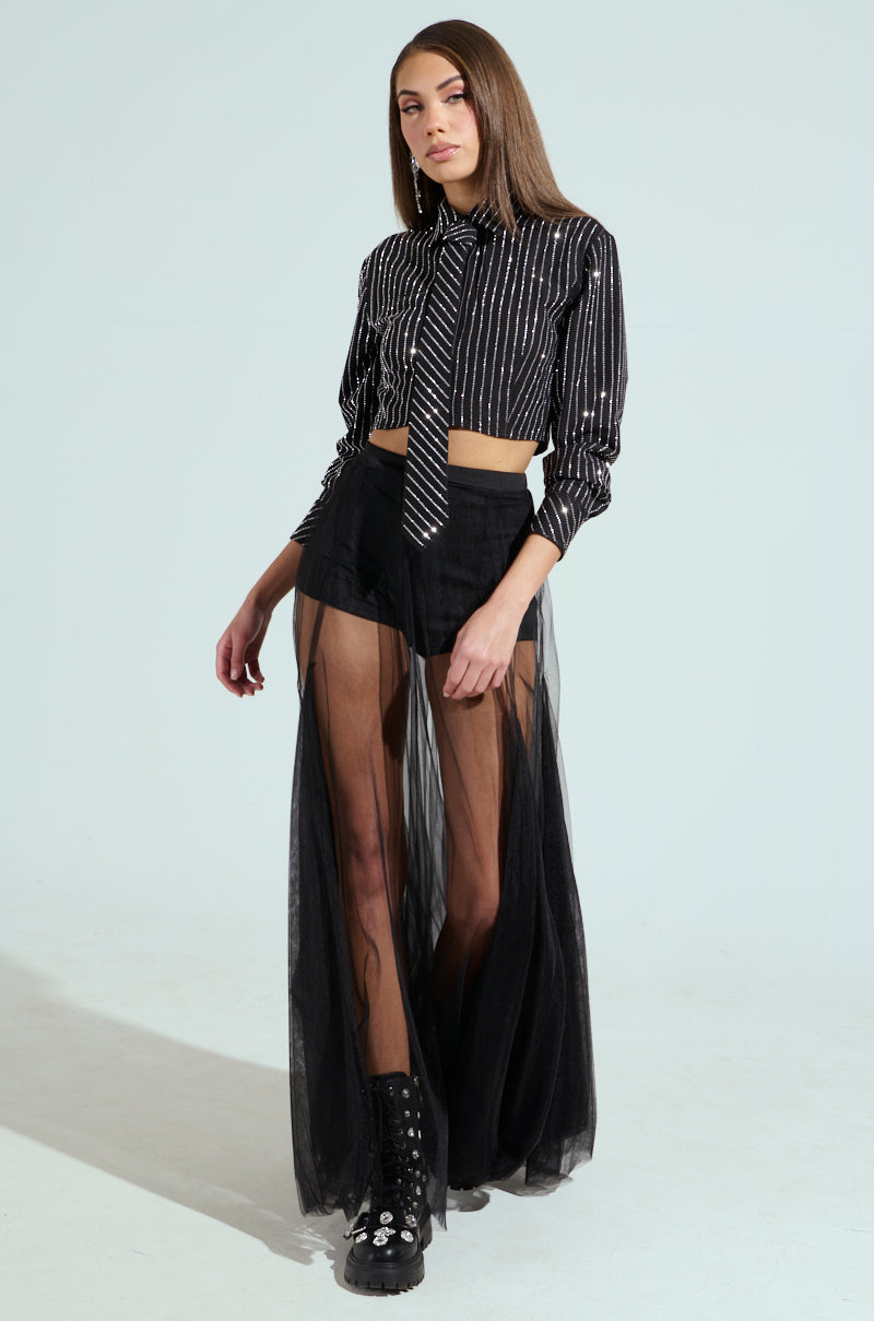A model poses in the "Just Go With It Button Down Top with Tie" paired with studded black boots. The boxy cropped silhouette enhances a sheer skirt over black shorts, crafting an edgy look against a plain backdrop.