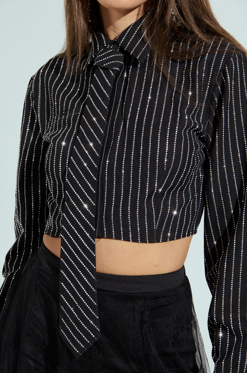 Dressed in the "JUST GO WITH IT BUTTON DOWN TOP WITH TIE," which is black with vertical white rhinestone pinstripes, a cropped boxy silhouette reveals the midriff. It's styled with high-waisted black pants against a light, plain background.