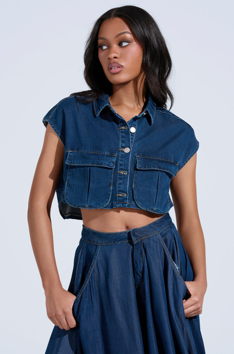 With long, dark hair, a person wears the CARRIE BUTTON DOWN DENIM VEST TOP, a sleeveless crop top with large pockets, paired with a matching high-waisted skirt. They gaze thoughtfully to the side against a plain background.