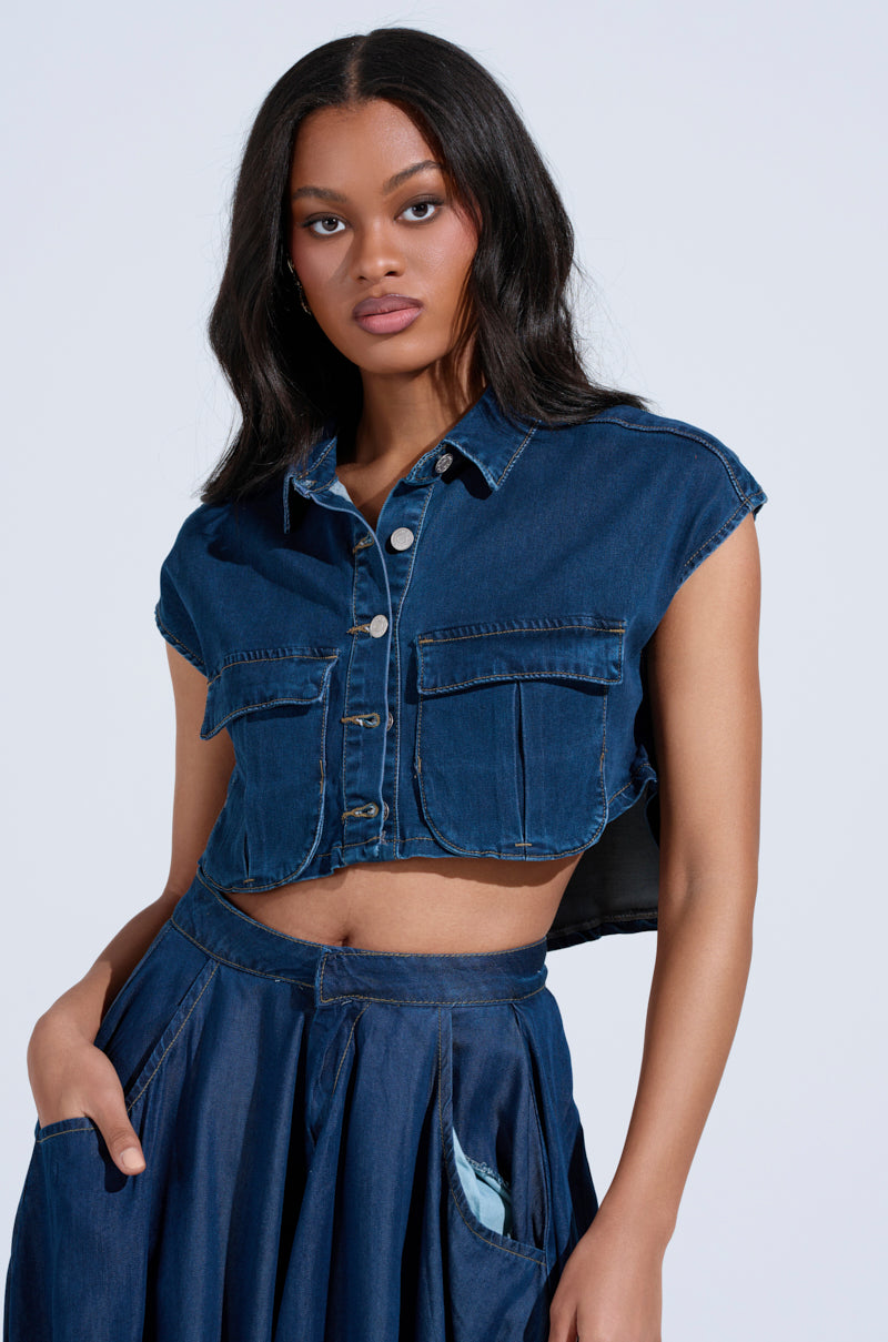 A person with long hair wears a sleeveless CARRIE BUTTON DOWN DENIM VEST TOP in a cropped silhouette, paired with a matching high-waisted skirt with small distressed details. The vest complements the outfit against the plain light gray background.