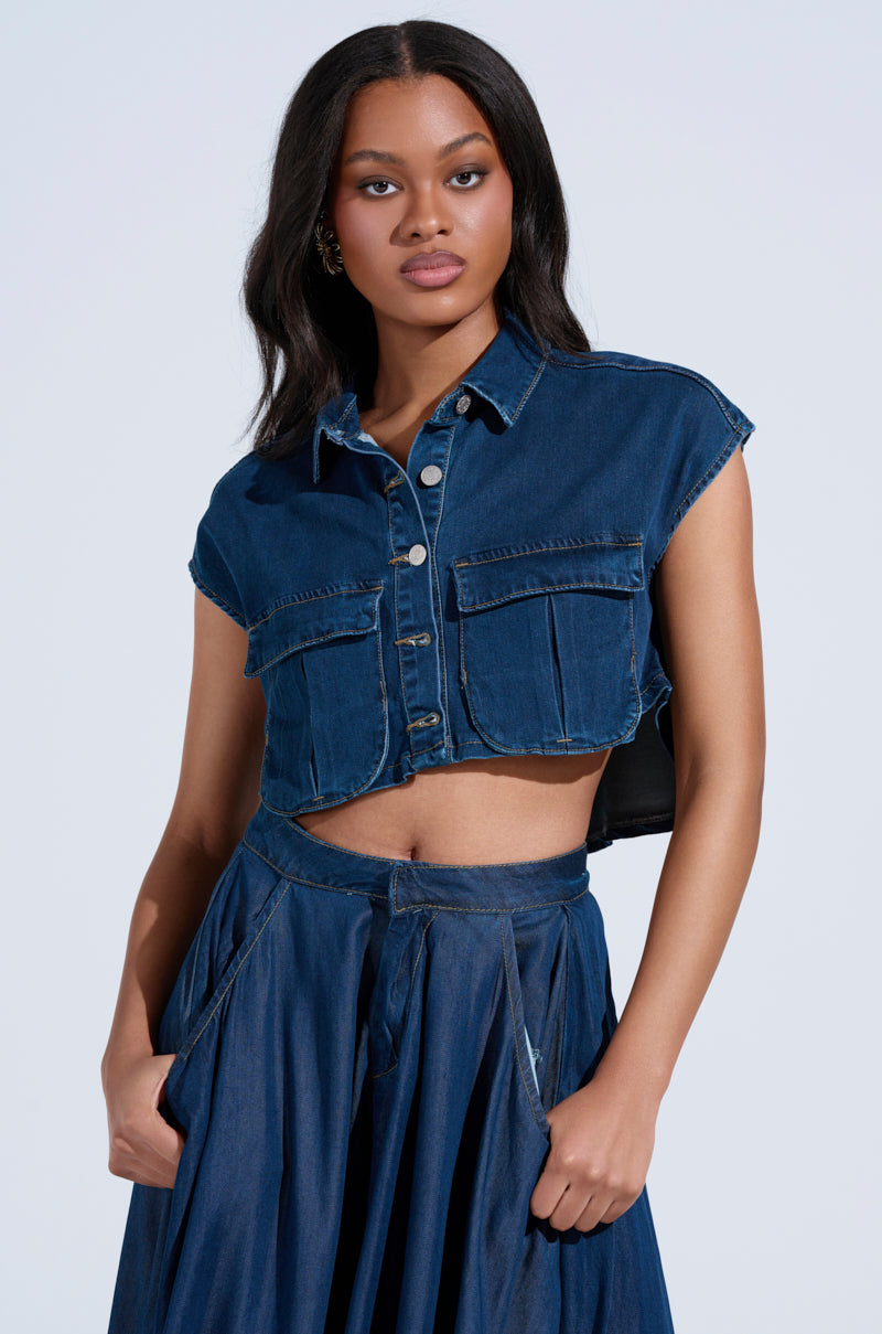 Wearing the CARRIE BUTTON DOWN DENIM VEST TOP with a high-waisted long denim skirt, someone stands against a neutral backdrop. The front button design and large pockets complement the cropped silhouette as their hands rest in the skirt's pockets.