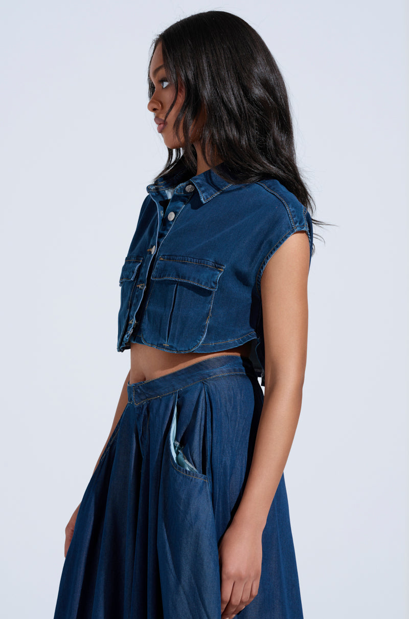 A person with long hair wears the CARRIE BUTTON DOWN DENIM VEST TOP, featuring a cropped silhouette and large pockets, paired with a high-waisted denim skirt. They stand in profile against a neutral background.