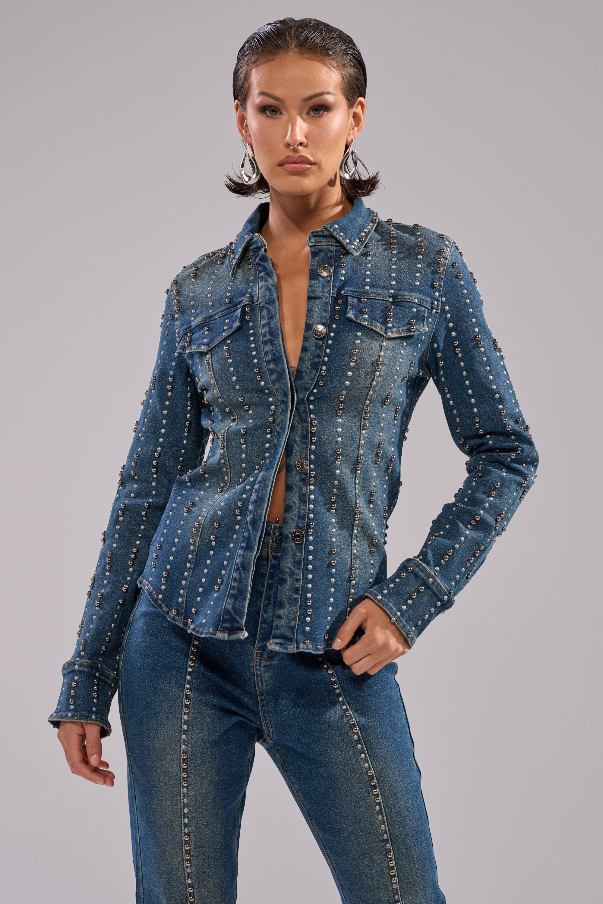A woman poses confidently against a plain gray background, wearing the GLAMOUR STUDDED BUTTON DOWN DENIM TOP with matching dark wash jeans.