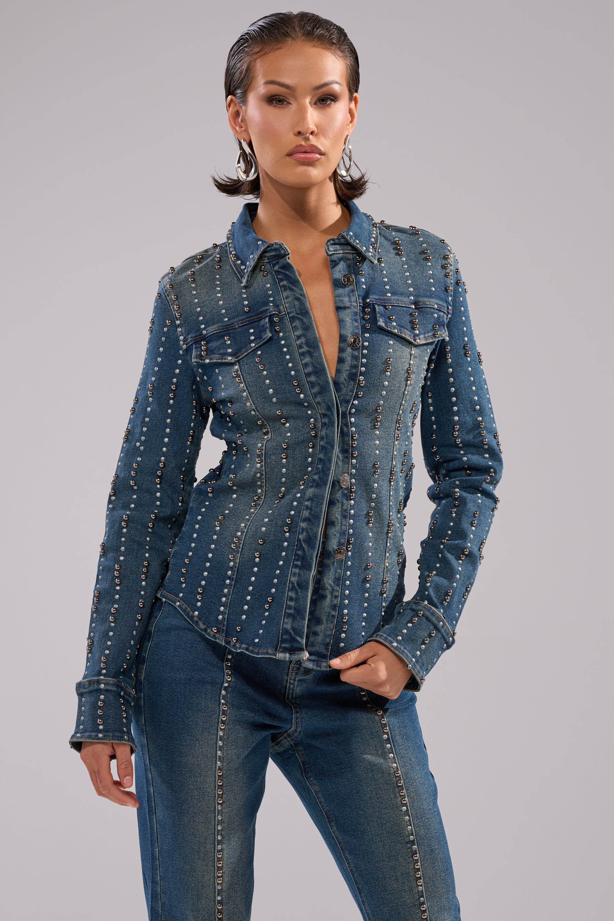 Wearing the GLAMOUR STUDDED BUTTON DOWN DENIM TOP with two chest pockets and matching dark wash jeans, a person stands confidently against a plain, light gray background.
