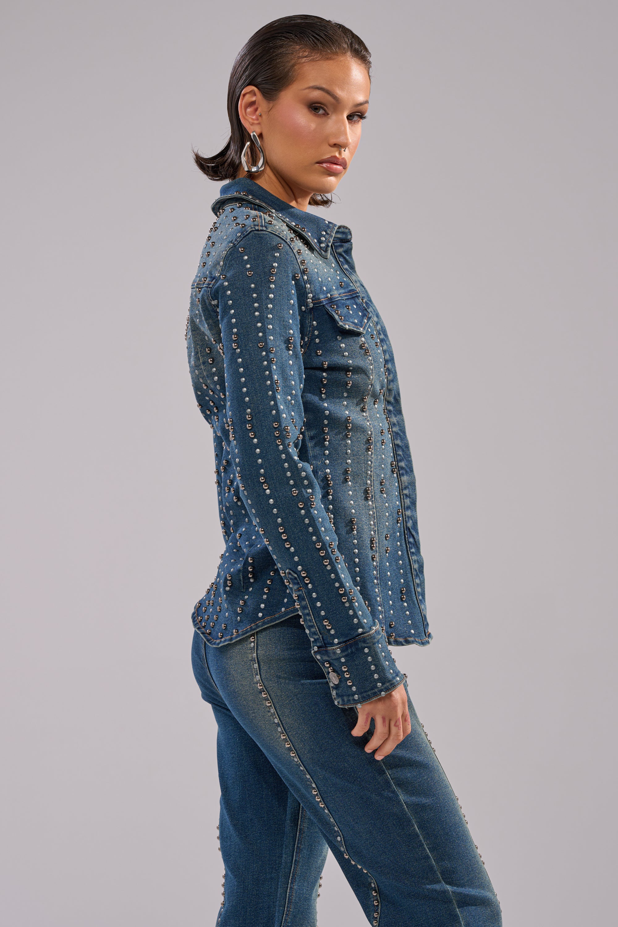 A woman in the GLAMOUR STUDDED BUTTON DOWN DENIM TOP and matching dark wash jeans stands in profile against a light gray background, styled with slicked-back hair and large hoop earrings.