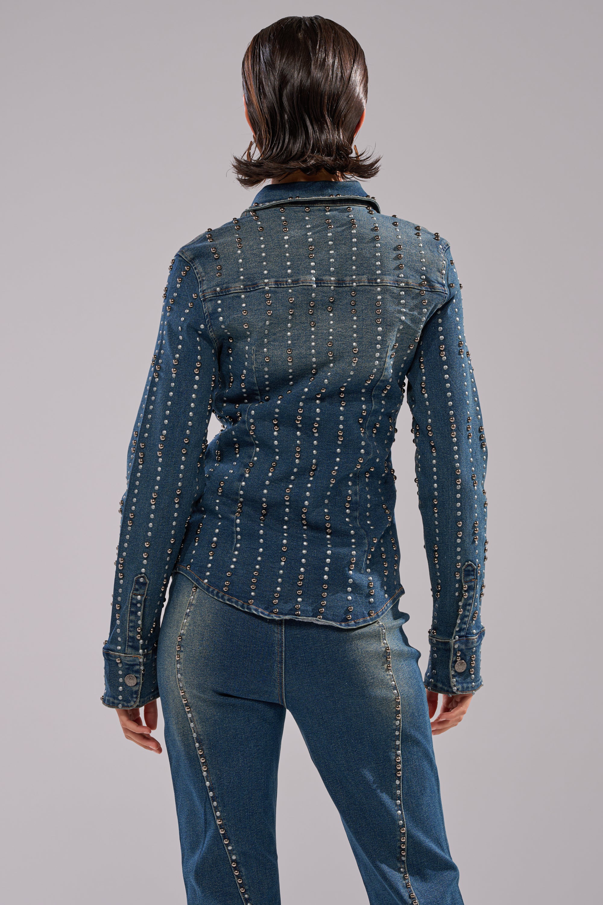 A person with short, slicked-back hair is seen from behind wearing the GLAMOUR STUDDED BUTTON DOWN DENIM TOP, featuring vertical metallic studs, paired with classic blue jeans.