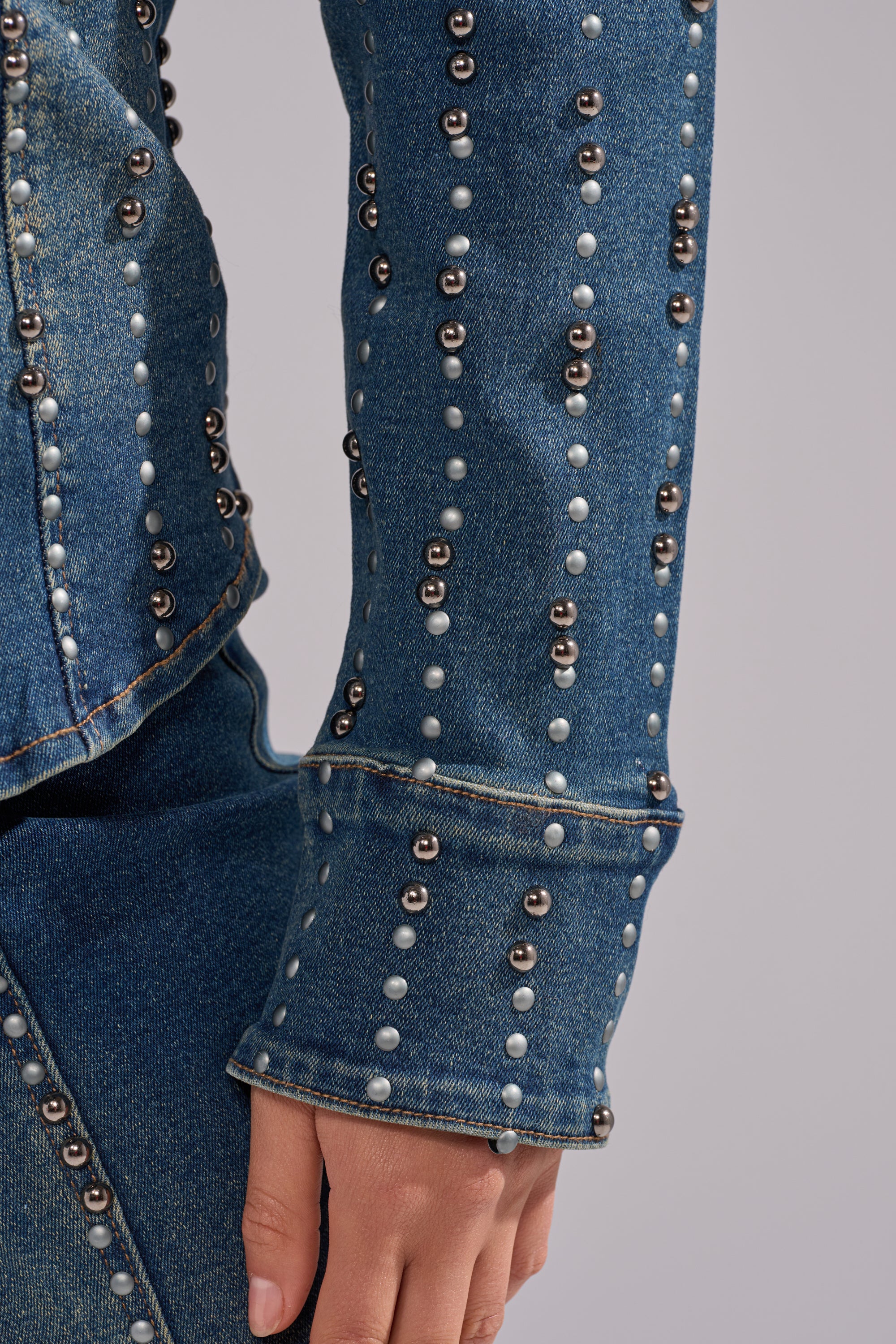 A close-up of a person’s arm and hand wearing the GLAMOUR STUDDED BUTTON DOWN DENIM TOP and jeans, both featuring vertical rows of metal studs and white pearls, set against a plain light grey background.