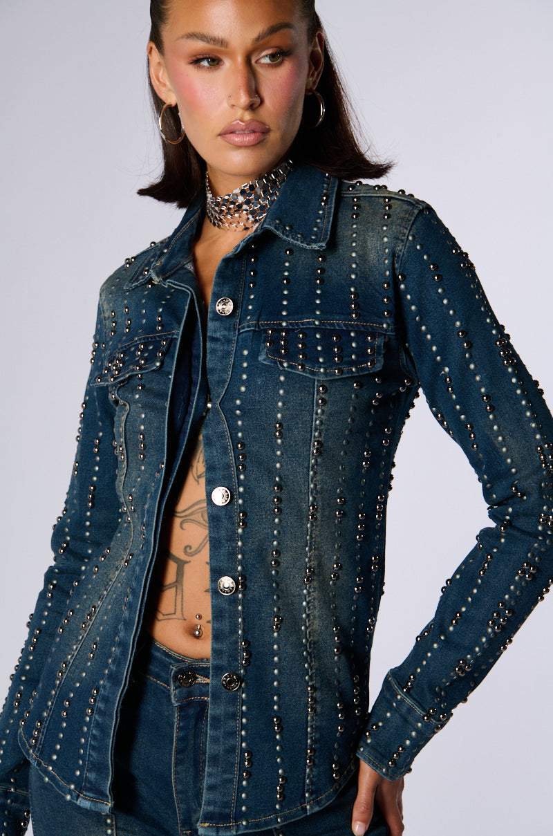 A person with long hair poses in JCR Trading LTD’s EVERYWHERE EVERYTHING BUTTON DOWN DENIM TOP, a dark denim jacket adorned with silver studs. They wear a matching studded choker and dark pants. The background is neutral, drawing focus to the outfit. 0