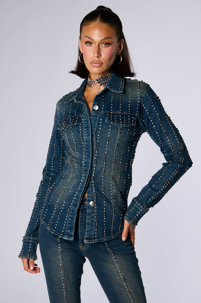 A person with long, dark hair is standing in a stylish pose, wearing the EVERYWHERE EVERYTHING BUTTON DOWN DENIM TOP by JCR Trading LTD. They accessorize with a choker necklace and large hoop earrings against a plain studio background, completing the look with matching denim jeans adorned with silver stud embellishments. 1