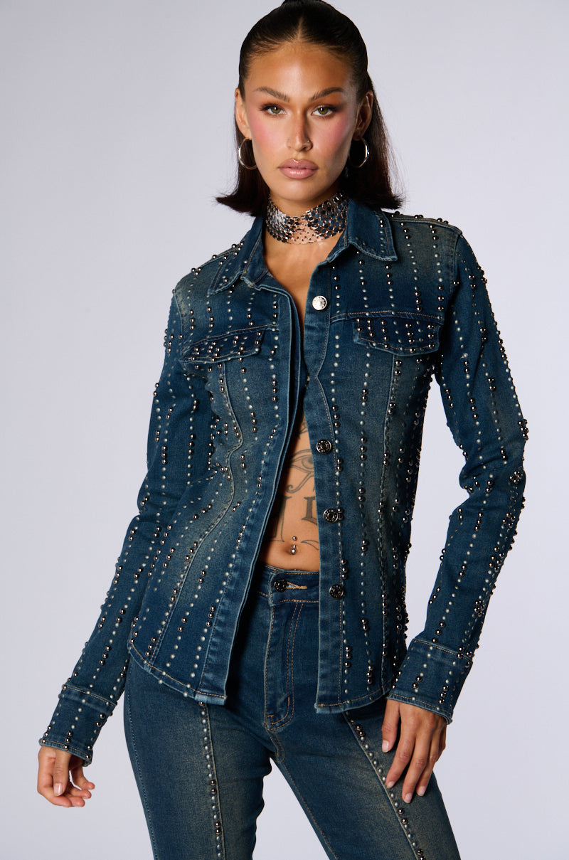 A person stands confidently in the EVERYWHERE EVERYTHING BUTTON DOWN DENIM TOP by JCR Trading LTD, paired with matching jeans. The denim top features vertical lines of metal studs. They wear a choker necklace and have tattoos visible on their torso. Their expression is serious, and their hair is styled back. 2