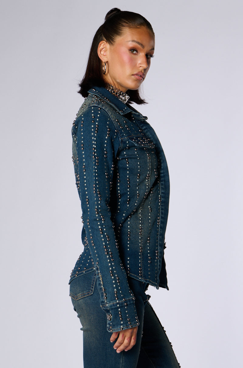 A woman with dark hair and a confident expression poses in the EVERYWHERE EVERYTHING BUTTON DOWN DENIM TOP by JCR Trading LTD, adorned with metallic studs. She stands in profile, looking over her shoulder towards the camera, also wearing matching studded denim pants against a plain background. 4