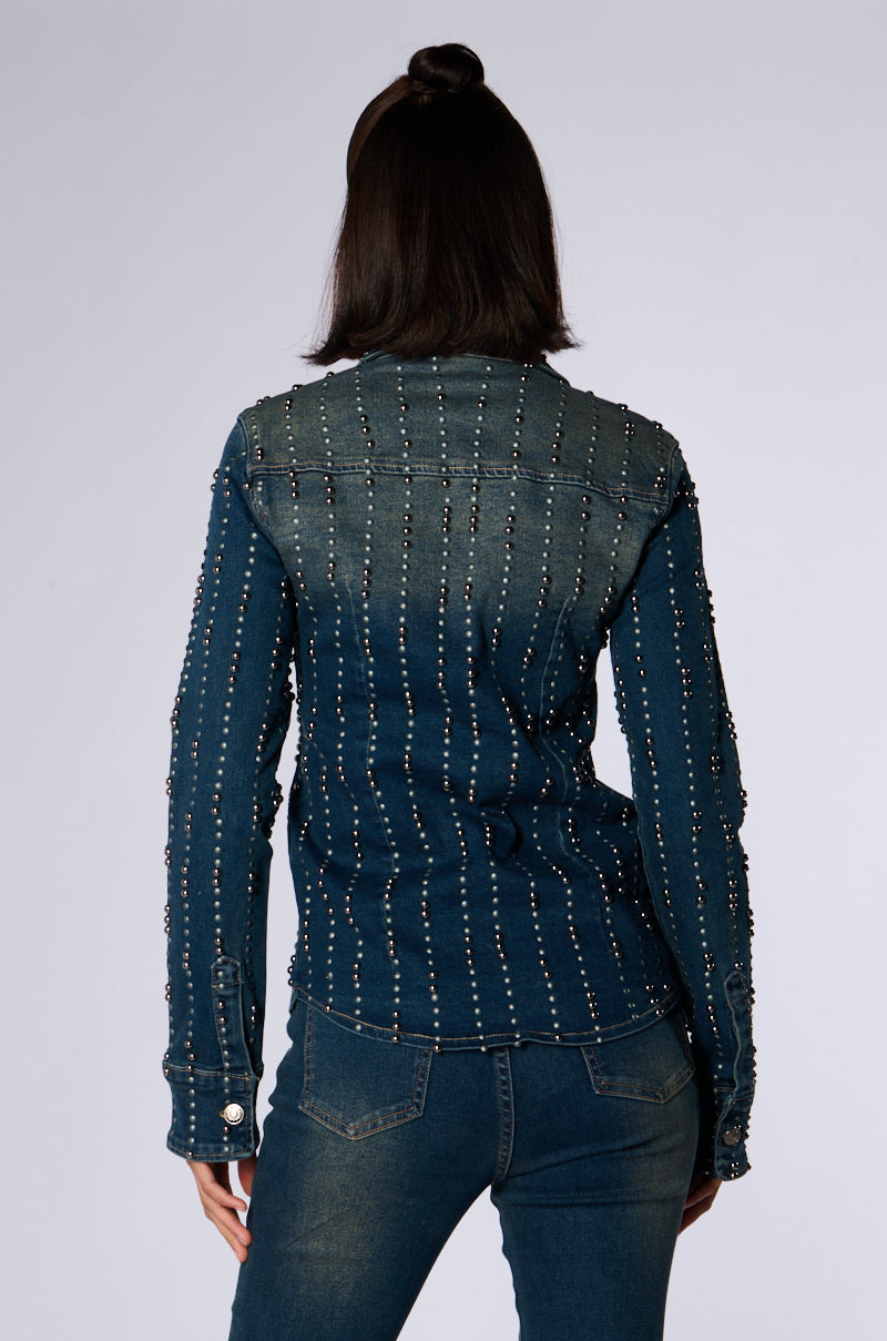 A person with shoulder-length dark hair, styled in a partial updo, is seen from the back wearing the EVERYWHERE EVERYTHING BUTTON DOWN DENIM TOP by JCR Trading LTD, adorned with vertical rows of small silver studs, paired with matching denim jeans. The background is plain and light gray. 5