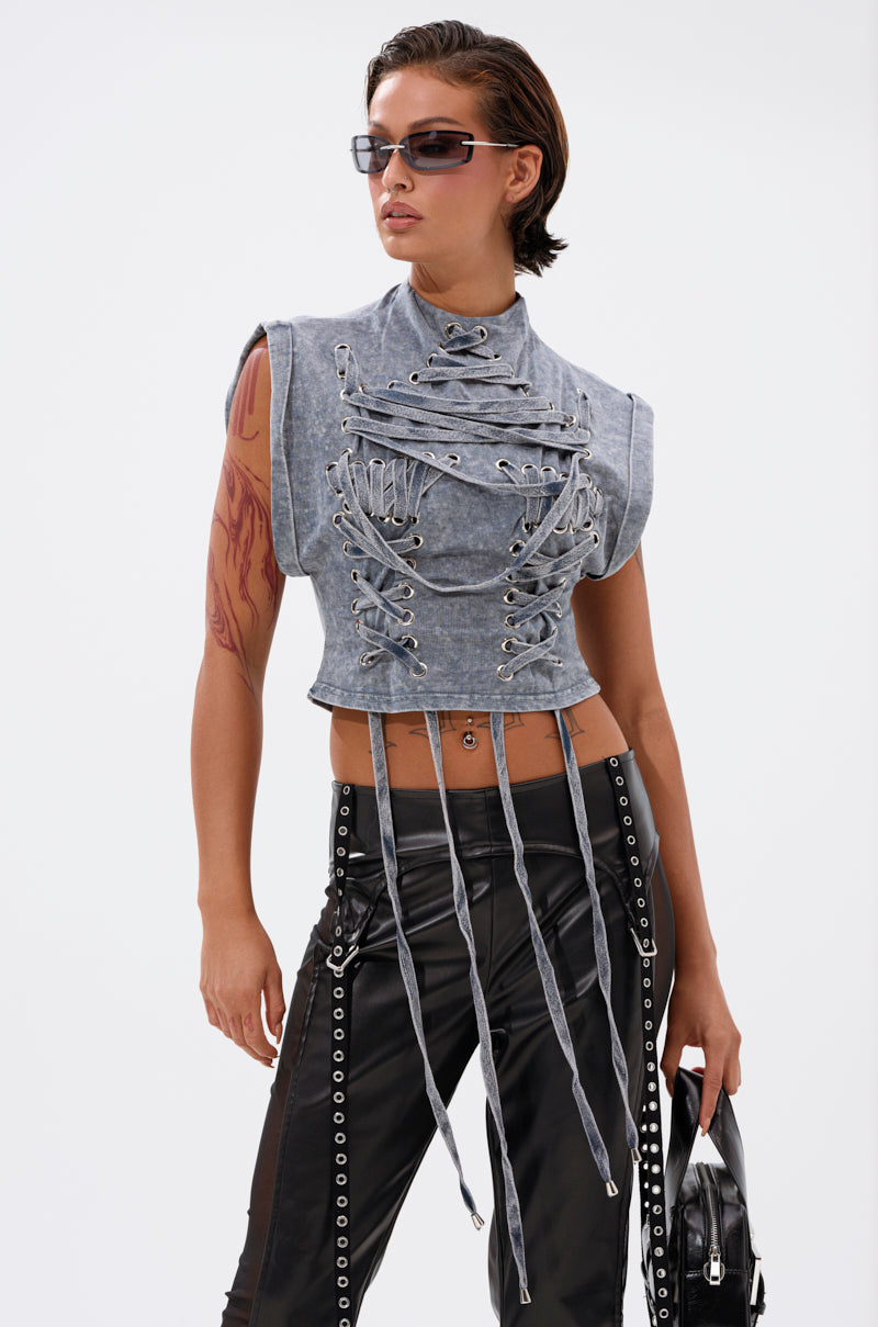 A person confidently stands against a plain white background wearing the ABLE TO SEE ME STRAPPY TOP, featuring a deconstructed corset-inspired gray design with hanging straps, paired with grommet-detailed black pants, sunglasses, and a black bag. 0