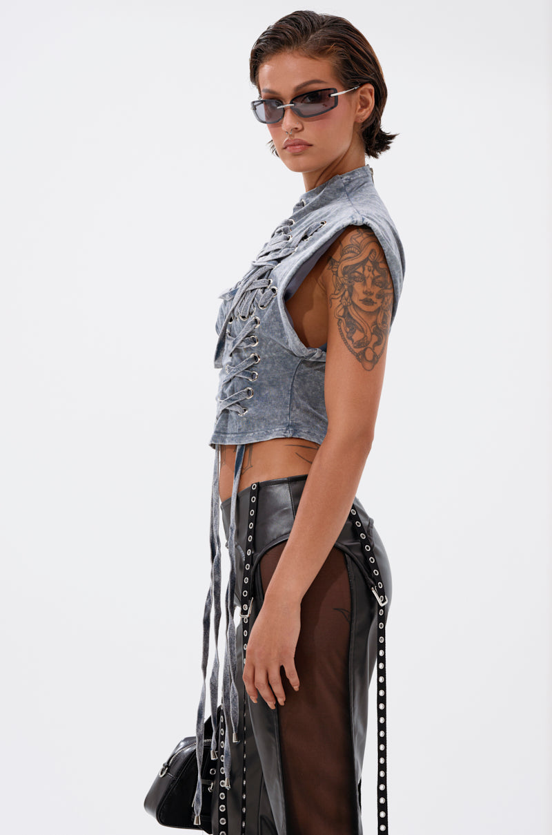 A person with short brown hair and sunglasses wears the ABLE TO SEE ME STRAPPY TOP and black leather chaps with sheer panels, showing a large upper arm tattoo, carrying a small black bag, and posing against a plain white background. 3