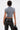 A person with short, wet-look hair is seen from behind wearing the ABLE TO SEE ME STRAPPY TOP in gray with cap sleeves and a zip-up front, paired with black high-waisted faux leather pants with strap details. Tattoos show as they hold a black handbag.