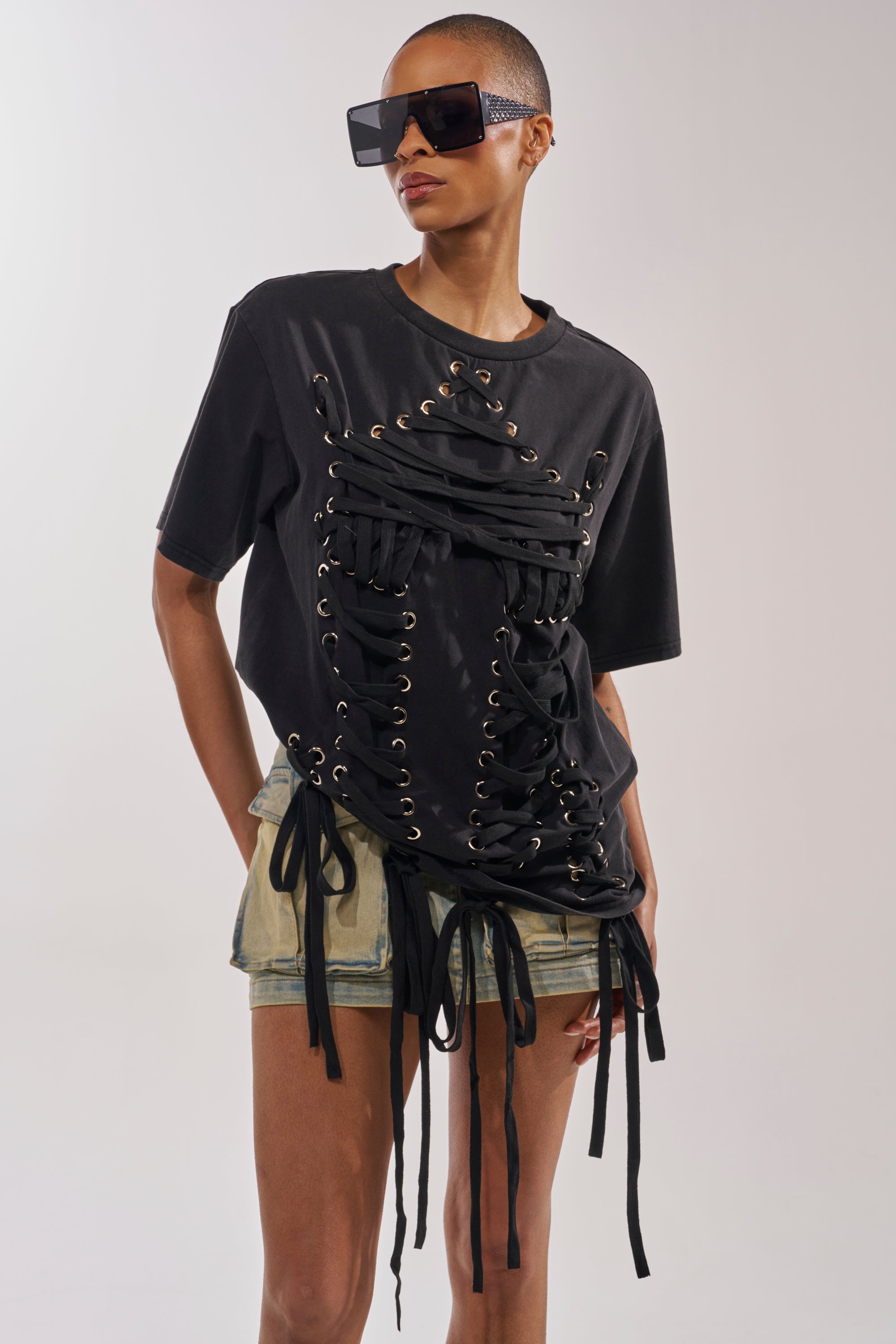 A person with short hair wears oversized black sunglasses, the LACED UP MINERAL WASH T-SHIRT with hanging ties, and light shorts, confidently standing against a plain background—a bold concert look. 0
