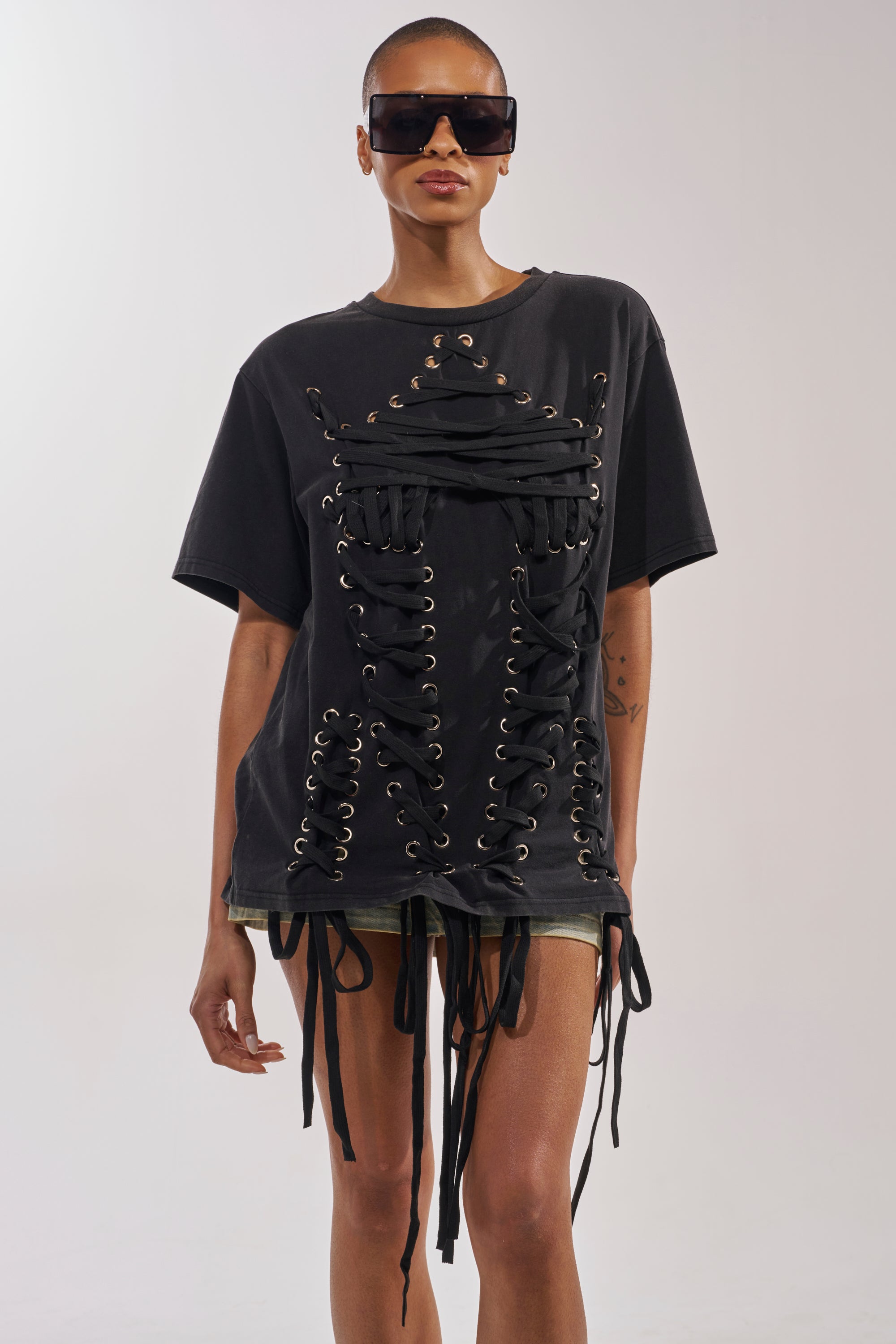 A person in oversized sunglasses and the LACED UP MINERAL WASH T-SHIRT with standout hanging ribbons poses against a plain light background. 1