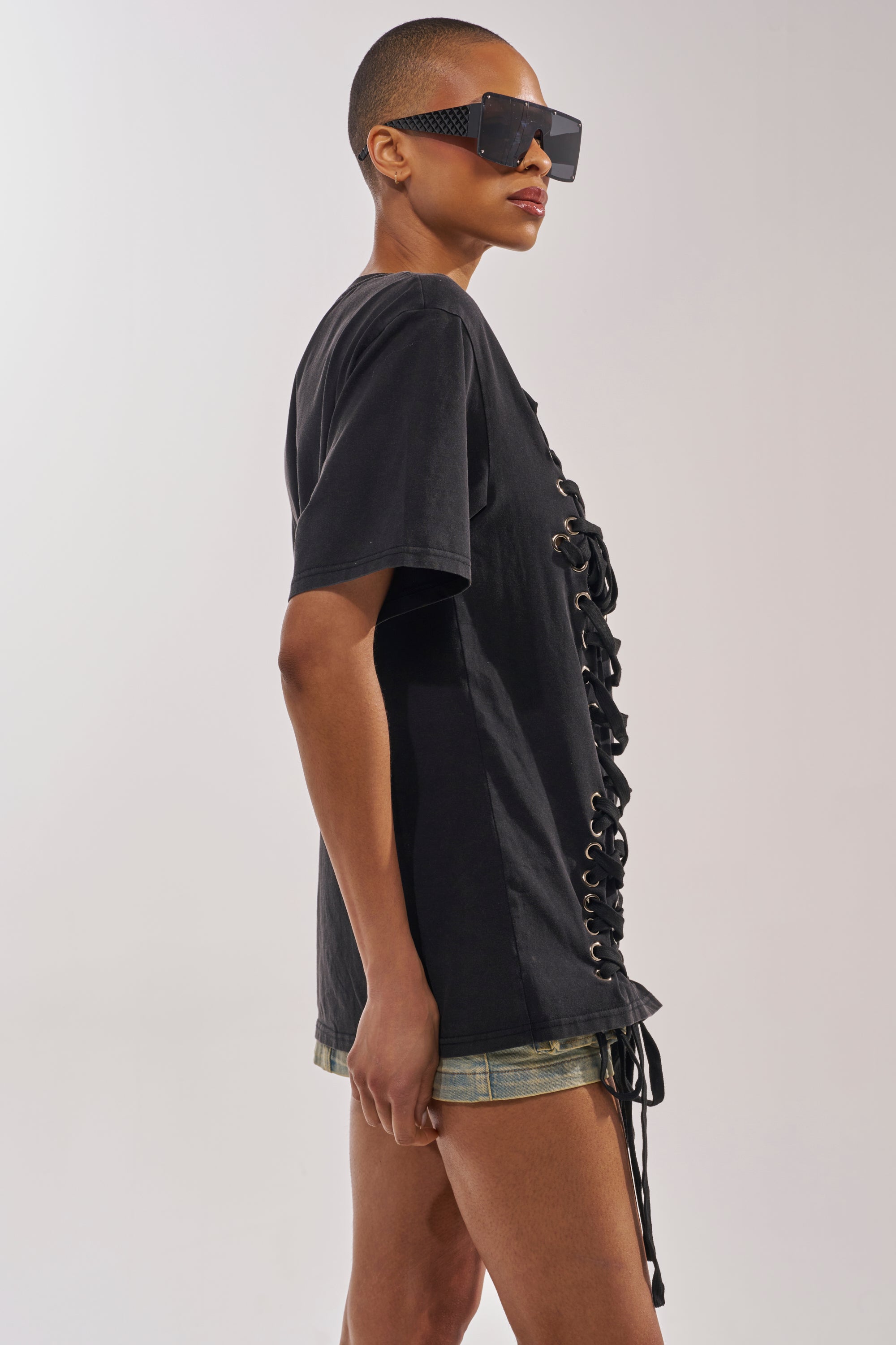 Wearing oversized black sunglasses and the LACED UP MINERAL WASH T-SHIRT with short denim shorts, a person stands sideways against a plain light background—a bold and stylish concert look. 2