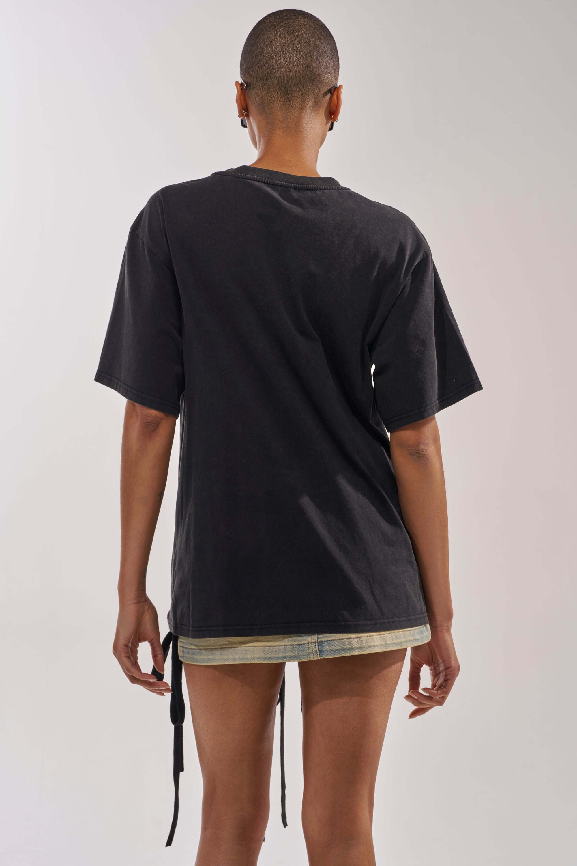 A person with short hair wears the LACED UP MINERAL WASH T-SHIRT and a short denim skirt, standing with their back to a plain light background. 3