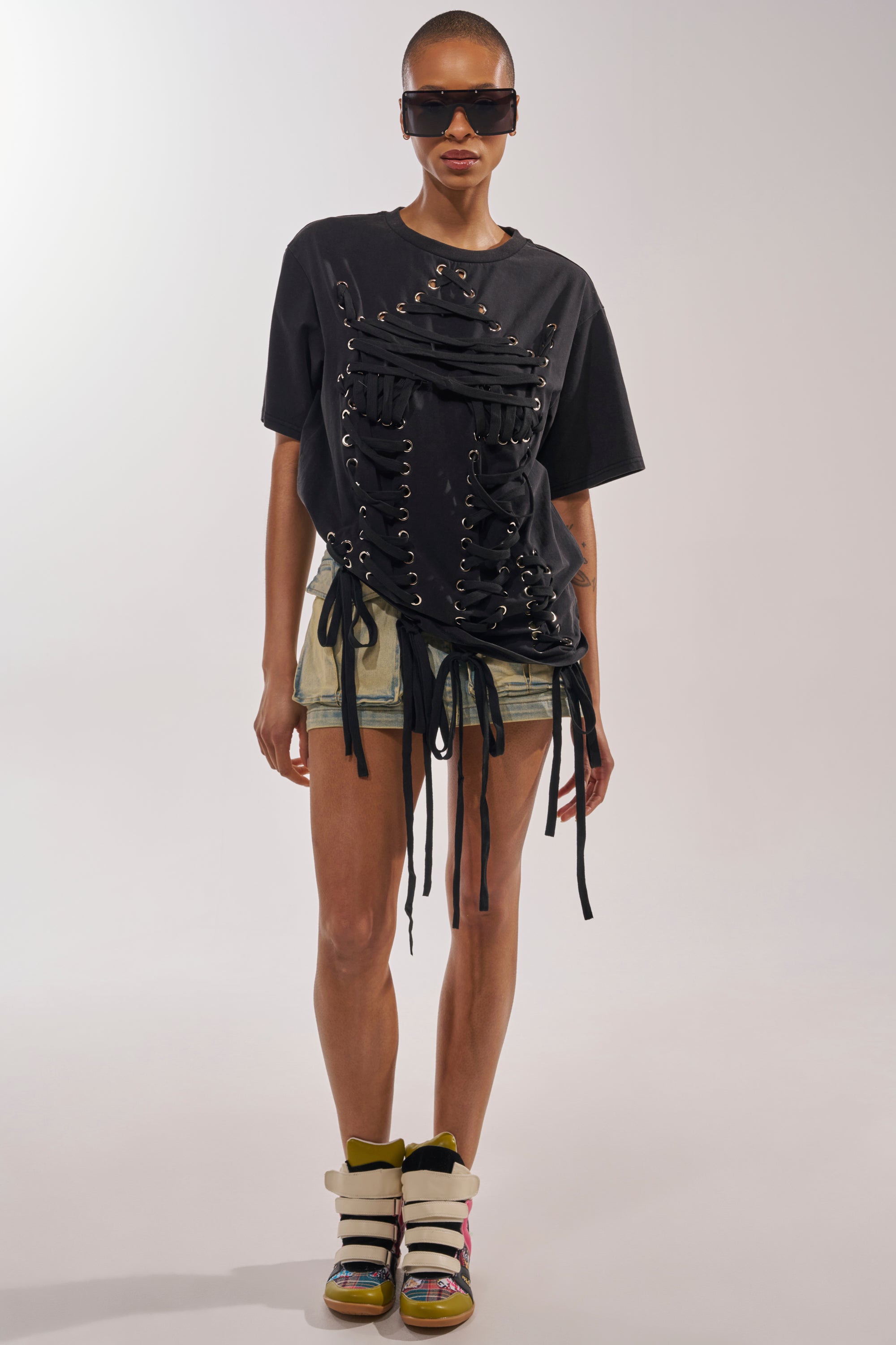 A person wearing sunglasses, a LACED UP MINERAL WASH T-SHIRT with grommet details, light denim shorts, and high-top sneakers stands against a plain light background—a cool concert look. 5