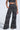 Wearing the "Set You Free Black Multi Palazzo Pant" set, featuring a black cropped top and high-rise, wide-leg pants with a swirling white chain pattern, they stand confidently with one hand in their pocket against a plain background.