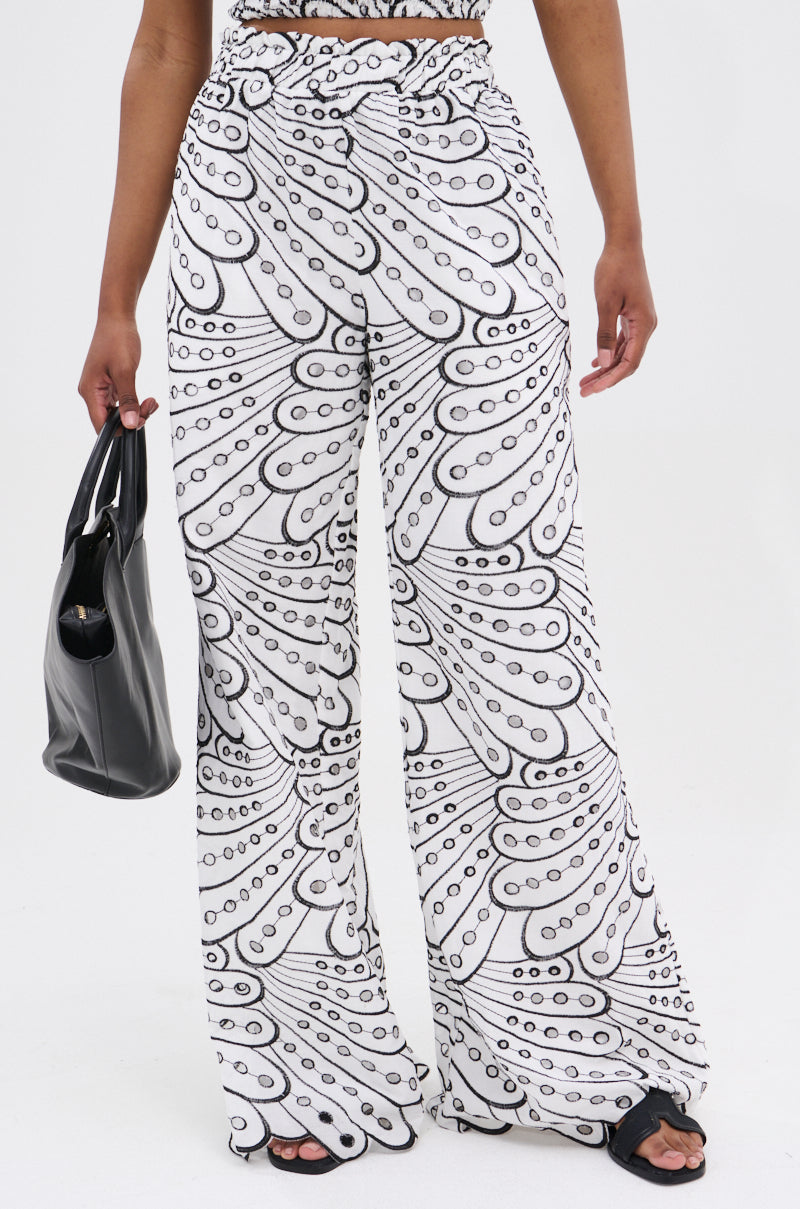A person models the AKIRA Label SET YOU FREE PALAZZO PANT—a high-rise, wide-leg white pant with a black abstract pattern—styled with black sandals and a black handbag, against a plain white background.