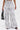 A person models the AKIRA Label SET YOU FREE PALAZZO PANT—a high-rise, wide-leg white pant with a black abstract pattern—styled with black sandals and a black handbag, against a plain white background.