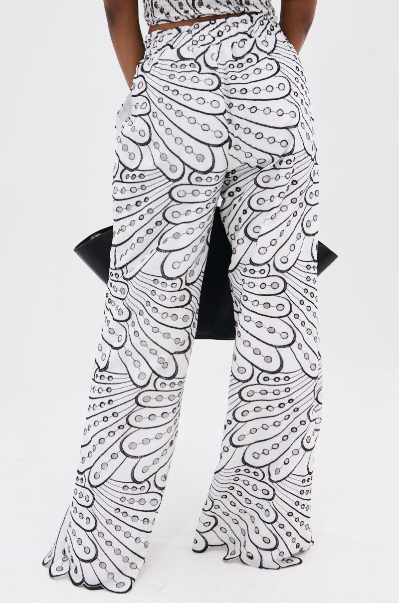 A person stands facing away, wearing the SET YOU FREE PALAZZO PANT by AKIRA Label, featuring a bold black and white abstract pattern. Their left hand is in a pocket, with a black clutch under their right arm. The background is plain white.