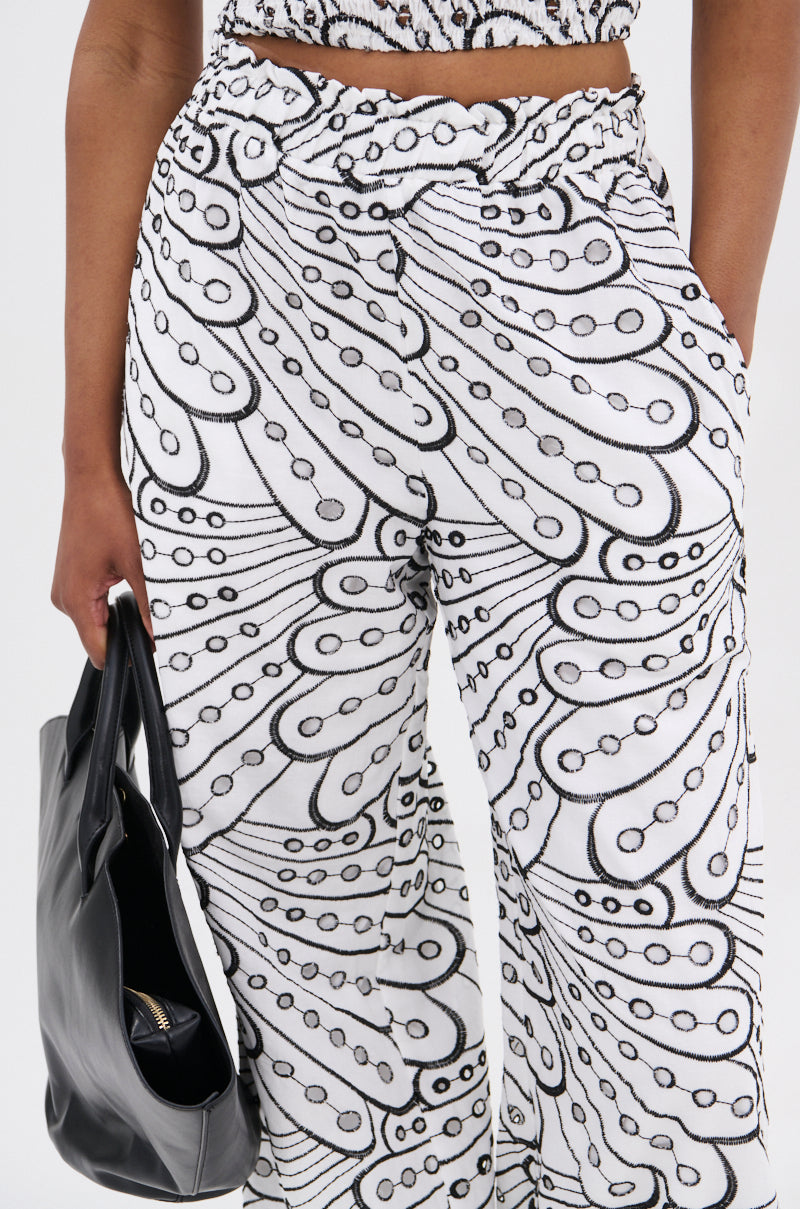 A person wears the SET YOU FREE PALAZZO PANT, a high rise white pant with bold black abstract swirls and dots, holding a black handbag. Only the lower torso and part of the hand are shown.