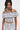 A woman with long dark hair in a half-up ponytail wears the SET YOU FREE OFF THE SHOULDER BLOUSE and matching pants with an abstract stitched pattern, standing against a plain white background and looking at the camera.