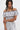 A woman with long, wavy black hair models the SET YOU FREE OFF THE SHOULDER BLOUSE with matching high-waisted pants featuring an abstract stitched pattern, standing against a plain white background.
