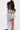 A woman with long, wavy black hair in a high ponytail models the SET YOU FREE OFF THE SHOULDER BLOUSE and matching skirt with an abstract stitched pattern, shown in profile against a plain white background for a cropped silhouette look.