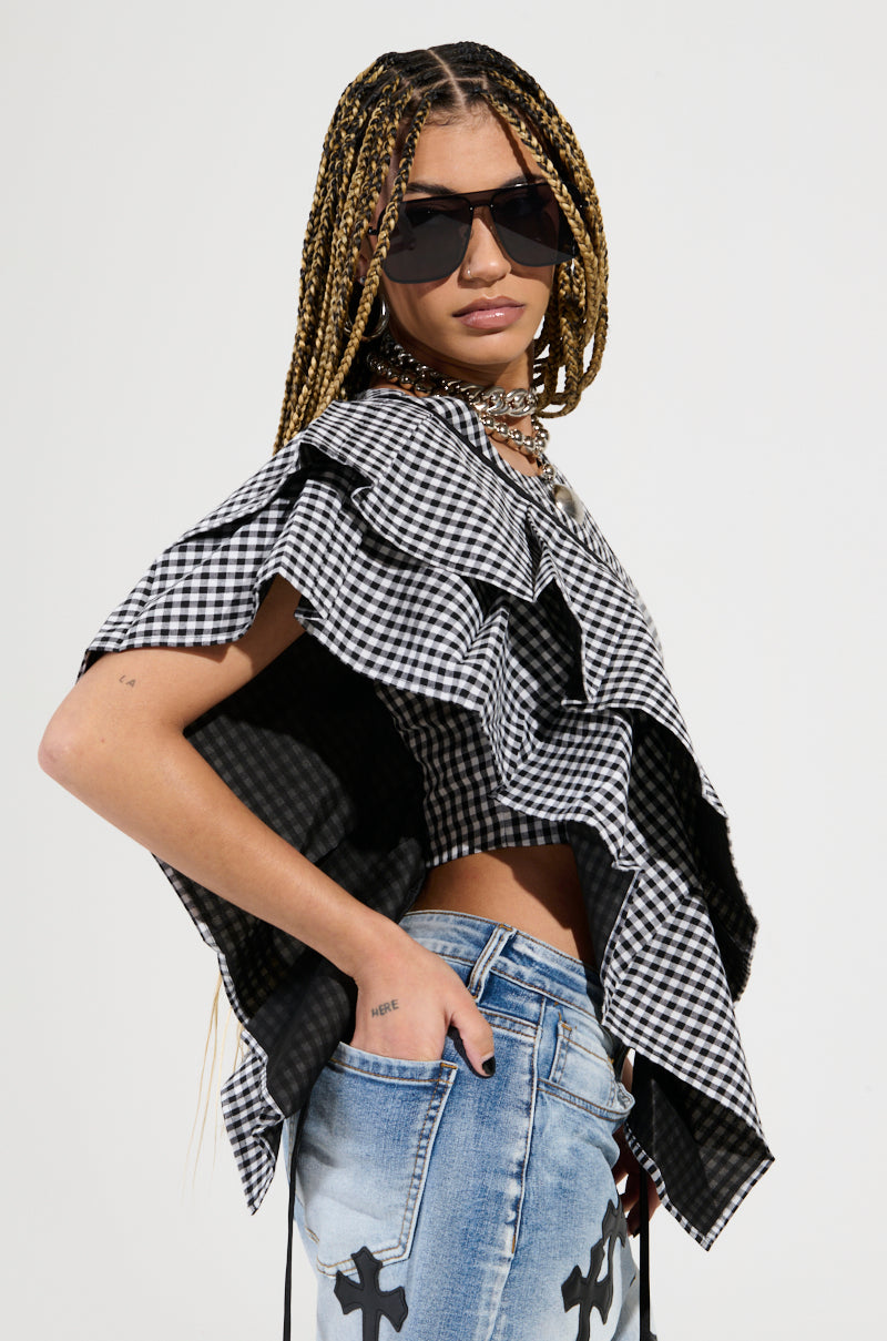 A person with long braided hair is dressed in an EASY ME GINGHAM BLOUSE featuring a black and white checkered pattern, paired with light blue jeans adorned with cross designs. They complement their look with dark sunglasses and a chunky necklace, confidently posing against a plain background.