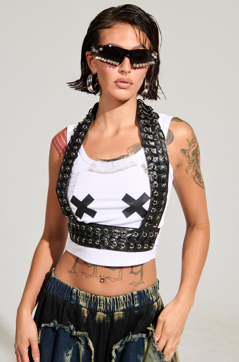 A woman with wet hair sports the LOCKED DOWN TANK, a white ribbed knit top highlighted by black Xs on the chest, complemented by a black leather harness. Her arms are adorned with tattoos, and she accessorizes like a true fashionista—donning dark sunglasses, silver earrings, and a denim skirt.