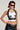 A fashionista with short dark hair rocks cat-eye sunglasses and the LOCKED DOWN TANK, a white ribbed knit crop top embellished with black Xs. Their studded leather vest highlights the tattoos on their arms and torso, while a dark denim skirt with a frayed hem adds an edgy touch. They pose confidently against a neutral background.